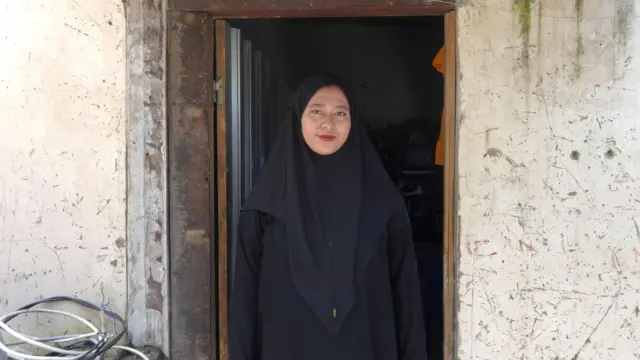 Siti Aminah berdiri di pintu, dengan jilbab dan baju warna hitam, tersenyum ke kamera.
