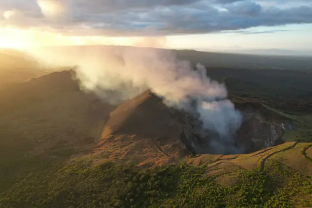 Una foto aérea del volcán Santiago, en Nicaragua. Se ve humo saliendo de su cráter. El humo va ascendiendo hasta que se confunde con las nubes en el cielo al atardecer.