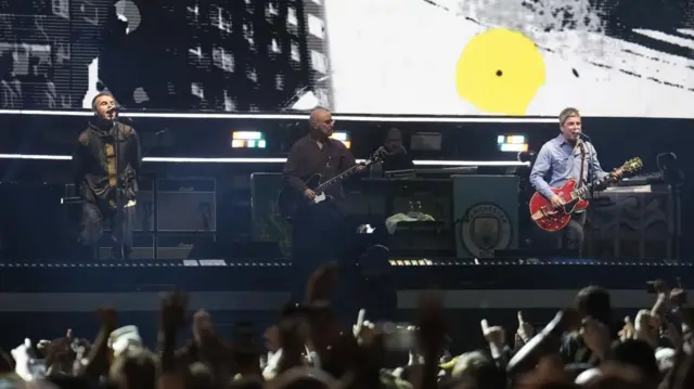 3人の男性がステージの上に並んで演奏している。向かって右側の男性は赤いギターを持ちながらマイクスタンドの前で歌っている。中央の男性は黒いギターを演奏している。左側の男性はカーキ色のジャケットのポケットに手を入れ、マイクスタンドの前で歌っている 手前にはコンサートに訪れた観客たちの後頭部が見える
Three members performing on stage at the Principality Stadium in Cardiff, Wales on 4 July. Concert-goers, some with their hands in the air, can be seen in the foreground.