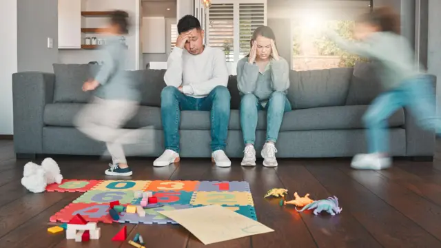 Ujęcie dwójki młodych rodziców wyglądających na zmęczonych, podczas gdy ich dzieci biegają po domu.
