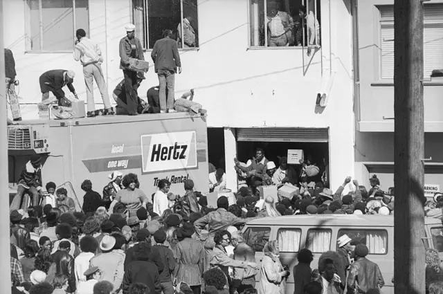Una calle en la que se están repartiendo alimentos, por parte del banco de alimentos de la familia Hearst. Hay mucha gente en la calle, en una escena desordenada.