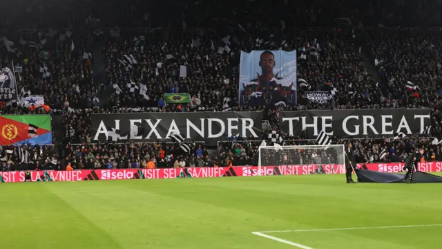 Les supporters de Newcastle United dans le Gallowgate End arborent un drapeau érythréen, une photo d'Alexander Isak et une pancarte en noir et blanc indiquant « Alexander the Great » (Alexandre le Grand) en référence à l'attaquant, alors que le terrain est pratiquement vide au premier plan.