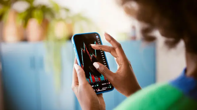 Mulher vista de costas com celular na mão, analisando gráficos de investimentos na tela do aparelho