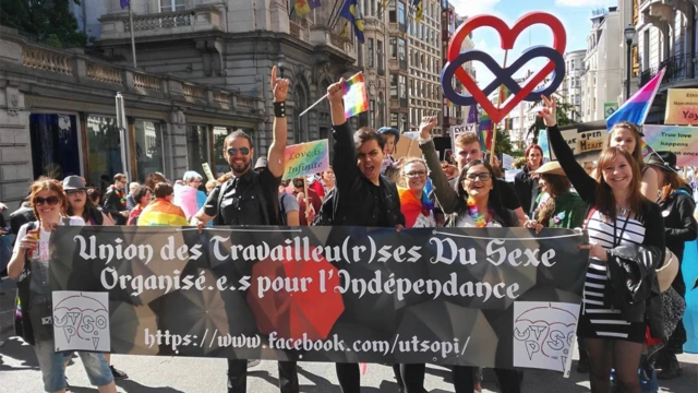 Une ligne de cinq personnes tenant une bannière noire, l'autre main en l'air, dans le cadre d'une manifestation - il y a beaucoup de monde derrière elles, dans une rue de la ville. La banderole représente l'Union belge des travailleurs du sexe (UTSOPI).