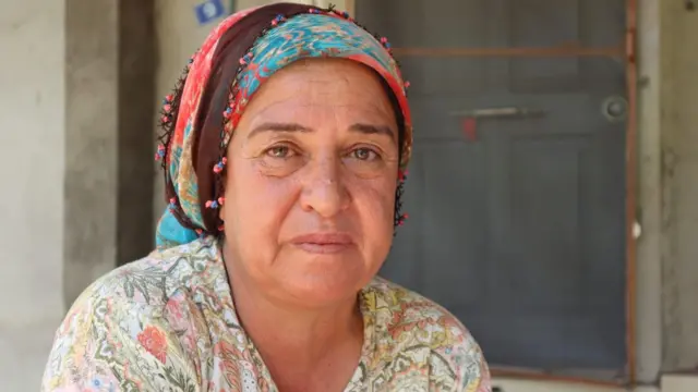 Renkli bir yemeni takmış orta yaşlı bir kadın fotoğraf çektiriyor. Yüzünde yorgun bir ifade var. Bir kapının önünde oturuyor.