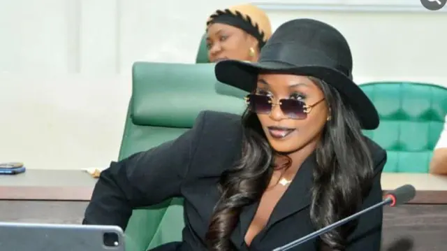 Ọnarebu Natasha Osawaru, o de fila dudu, wọ aṣo dudu o si lo igo dudu pẹlu. O n rẹrin-in musẹ labẹ igo rẹ, ẹrọ gbohun-gbohun kan wa niwaju rẹ pẹlu. Irun didan alawọ dudu to gun walẹ lo ṣe ninu aworan yii