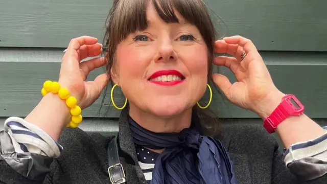 Caroline Jones está de pie sobre un fondo de madera verde pálido, mirando a la cámara y sonriendo. Tiene cabello castaño con flequillo, ojos azules y lleva un labial rojo brillante. Tiene los brazos levantados y se toca las orejas. Se ve la parte superior de una chaqueta gris oscura, así como una bufanda azul y una blusa de rayas azules y blancas. También lleva una pulsera de cuentas amarillas, pendientes de aro amarillos y un reloj rosa.