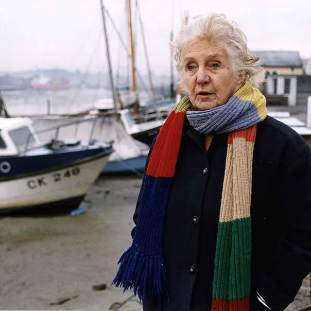 Joan Hickson cerca de un muelle, viste un abrigo azul y una bufanda de colores. Es una persona de mayor edad y tiene el cabello canoso. 