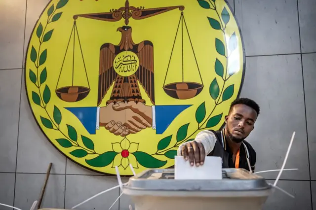 Homem segurando uma cédula de papel que ele deposita em uma urna, com o emblema nacional da Somalilândia ao fundo. O emblema é redondo e amarelo, com o desenho de duas mãos se cumprimentando no meio, além de uma ave e uma balança representando a Justiça