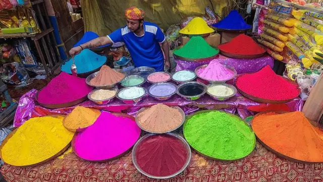 Muškarac na pijačnoj tezgi okružen metalnim činijama ispunjenim raznim bojama