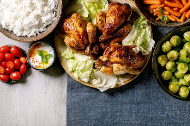 Table de dîner avec mini-poulet grillé, riz et légumes choux de Bruxelles cuits au four, carottes miniatures dans des bols en céramique sur une nappe en lin bleu sur fond gris.
