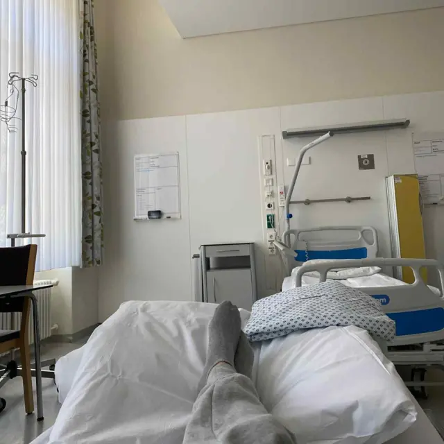 Людина лежить у лікарняному ліжку, видно її ноги в сірому одязі й шкарпетках. Перед нею – простора, світла палата з білими стінами, сучасним обладнанням, ще одним порожнім ліжком, тумбочкою та вікном із напівпрозорими шторами.