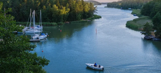 Rzeka płynie spokojnie przez lesisty krajobraz, tworząc zakola i rozlewiska. Po lewej stronie widać kilka zacumowanych żaglówek, na pierwszym planie płynąca motorówka. 