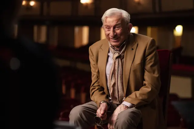 Haldun Dormen sahnede oturmuş, elinde gözlük, gülümsüyor. Üzerinde açık kahverengi bir ceket ve fular var