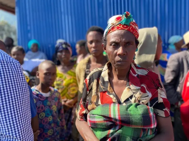 Umugore wambaye ishati y'amabara arimo umutuku, umweru n'umukara ahagaze areba muri 'camera' ateze igitambaro mu mutwe kirimo amabara y'icyatsi kibisi, umutuku n'umweru. Acyenyereye mu gatuza undi mwitero urimo amabara y'icyatsi kibisi n'umutuku. Akikijwe n'abandi bantu, inyuma yabo hari inyubako y'amabati y'ubururu. Ifoto yo ku itariki ya 29 Kanama (8) mu 2025. 