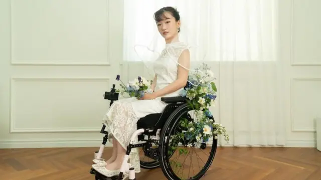 한 여성이 흰 웨딩드레스를 입고 휠체어에 앉아 있다. 휠체어에는 초록색, 흰색, 파란색 꽃들이 장식되어 있다.