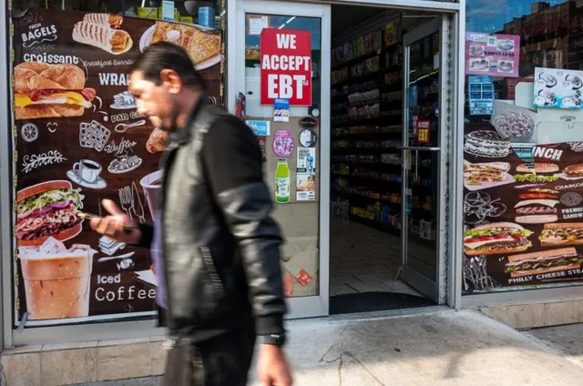 تابلویی با نوشته «EBT قبول می‌کنیم» (کارت کمک‌های اجتماعی) ، بیرون یک فروشگاه مواد غذایی در بروکلین نیویورک نصب شده است، عابری در حال گذر از مقابل فروشگاه است، ششم نوامبر ۲۰۲۵