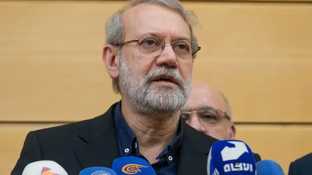 Siyah bir ceket, mavi açık yakalı düğmeli bir gömlek ve gözlük takan Tuğgeneral Ali Larijani, 27 Eylül 2025'te düzenlenen bir anma töreninde mikrofonların önünde konuşuyor.