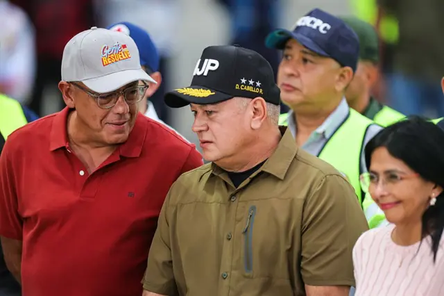 Jorge Rodríguez, a la izquierda, vestido con oolo rojo y gorrra gris, parece decirle algo a a Diosdado Cabello, en el centro, con camisa color militar y gorra negra y, en la esquina a la derecha, Delcy Rodríguez, que sonríe. 