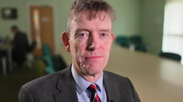 Jonathan Hall, secretario del Consejo Parroquial de Hellesdon, mira a la cámara. Tiene el pelo castaño claro y ralo, viste traje oscuro, camisa azul claro y corbata a rayas rojas, azules y doradas.