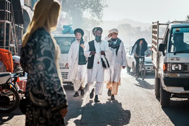 Taliban morality police patrol the streets dressed in white to enforce dress attire. Woman photographed wearing yellow headscarf, crossing the street.