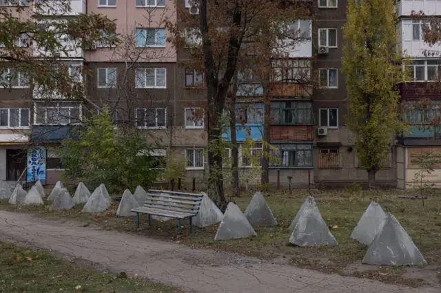 Подвір’я біля багатоповерхового житлового будинку. Уздовж стежки стоїть ряд бетонних пірамідальних протитанкових укріплень, поруч – стара металева лавка. Дерева навколо частково облистянілі, а будинок має потертий фасад із різнокольоровими балконами.