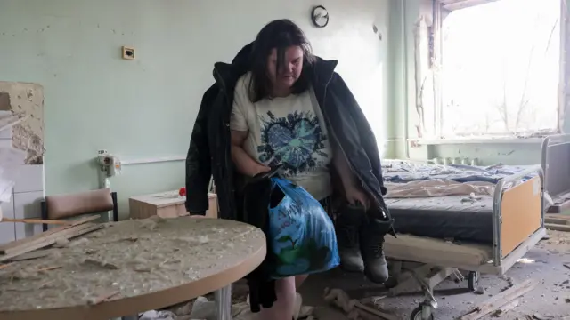 産科病院の病室で、女性が大きな黒いコートを肩にかけ、片手に衣類が詰まった青いビニール袋を、もう片手にブーツを持っている。背後の窓は吹き飛ばされ、割れたガラス片やがれきが床、ベッド、その横のテーブル一面に散乱している。