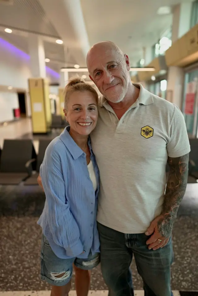 Dave Green e Andrea Urmston, um homem e uma mulher de meia idade, sorriem e olham para a câmera. Eles estão próximos um ao outro e ela é mais baixa do quele. Ela veste camisa azul clara sobre uma camiseta branca, e bermuda jeans rasgada nas coxas. Ele veste camisa polo cinza e calça jeans