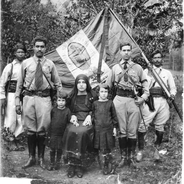 Una mujer vestida de negro junto a dos niños y rodeada de soldados con la bandera de México de espaldas en la que se ve una imagen de la Virgen de Guadalupe.