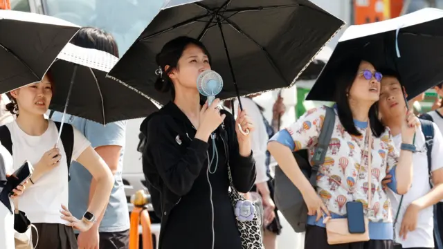 Tri Japanke sa kišobranima, među kojima jedna pokušava da se rashladi malim ventilatorom