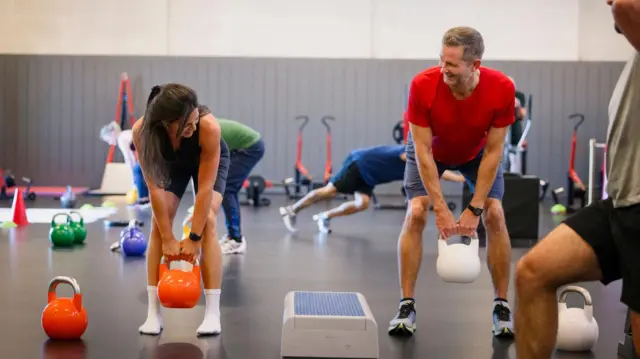 Mujer joven levantando una pesa rusa de color naranja mientras entrena con una amigo en el gimnasio