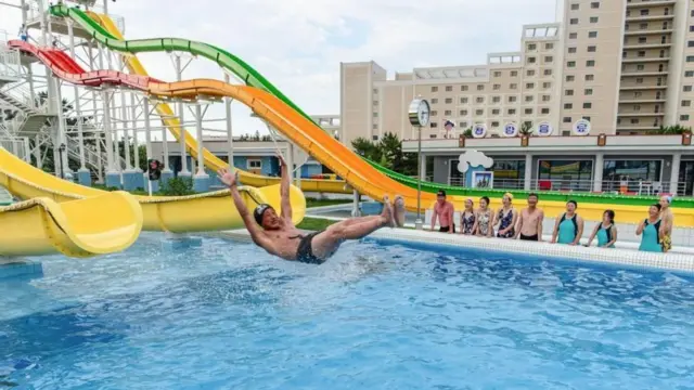 Un hombre cayendo en una piscina tras deslizarse por un tobogán amarillo gigante mientras otros nadadores lo observan emocionados