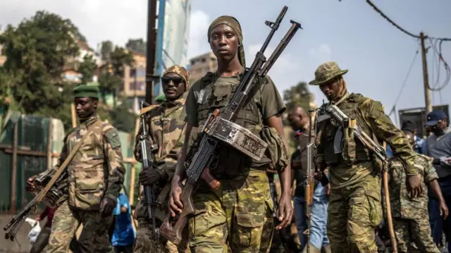 Un groupe de combattants du M23, vêtus de treillis militaires, portent des armes lourdes.