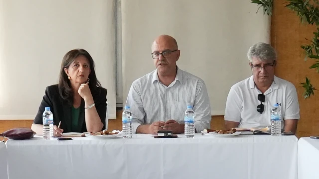 siyah resmi kıyafetli bir kadın, kel ve gözlüklü bir adam ve yanında beyaz saçlı gözlüklü bir erkek beyaz örtülü bir masada oturuyor.