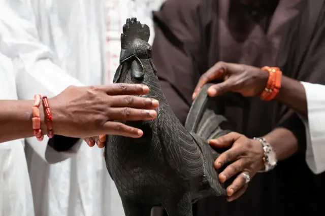 Nigerian officials place their hands on the bronze cockerel.