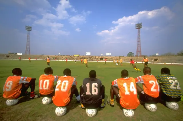 Un groupe de jeunes footballeurs assis sur des ballons de football regardent un match d'essai qui se déroule dans le stade national de Lusaka.