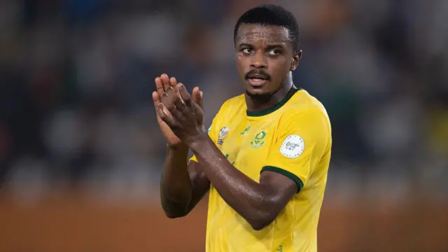 Teboho Mokoena, wey dem picture from waist up for di traditional South African gold jersey, clap im hands as dem sub am off during one football match