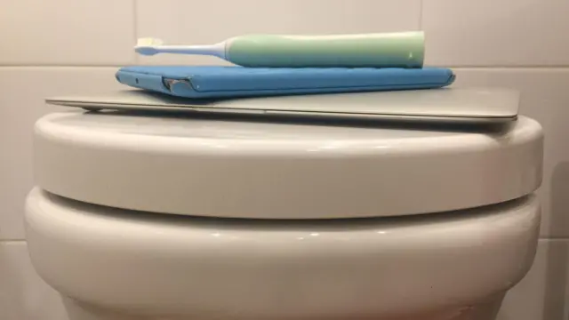 Uma escova de dentre em cima de um tablet e um computador que estão em cima de um vaso sanitário com a tampa fechada