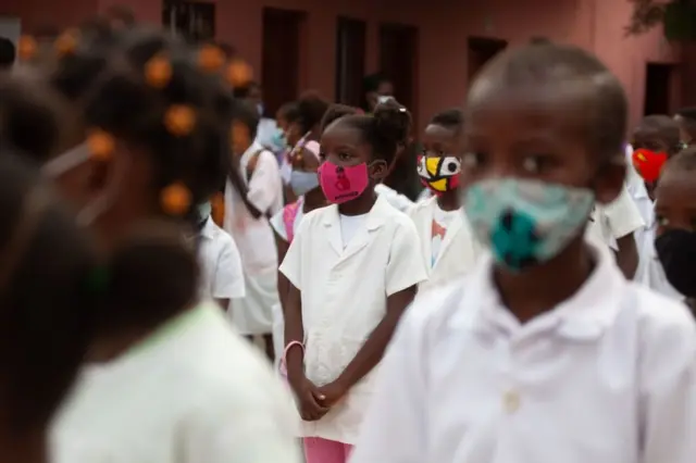 Primary school students wear masks and uniforms as they wait in a courtyard before lessons begin.