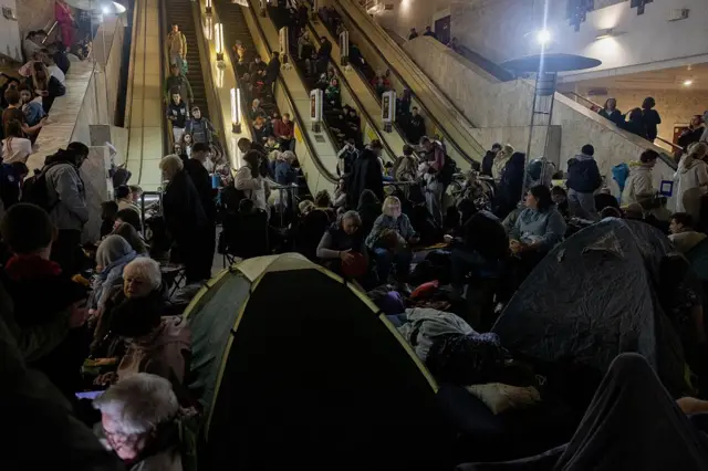 Багато людей перебувають у приміщенні станції метро: сидять на підлозі та сходах, поруч із речами й наметами, використовуючи простір як укриття.