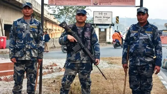 पूर्वी सीमाना मेचीपूलमा तैनाथ शसस्त्र प्रहरीहरू। तीन जनामध्ये एक जना प्रहरी अधिकृतले बन्दुक समाइरहेका छन् भने मेचीपुल लेखेको बोर्डको मुन्तिर एउटा स्कुटरमा सवार दुई जना महिलाहरू देखिन्छन्