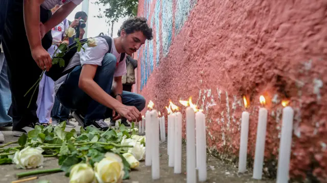 Homem branco acende velacomo ganhar dinheiro com palpites de futebolfrente a um murocomo ganhar dinheiro com palpites de futebolescola, há rosas no chão