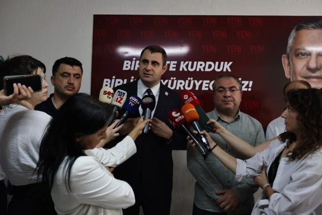 Yeni Parti İzmir İl Başkanı Çağatay Güç gazetecilere açıklama yapıyor. Gazeteciler ellerinde mikrofon tutuyor. Çağatay Güç'ün yanında iki adam var. Arkadaki posterde ise Özgür Özel görülüyor.