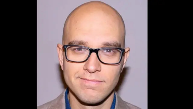 Une photo de tête du professeur Sébastien Jodoin. Il est chauve, porte des lunettes noires et sourit à l'appareil photo.