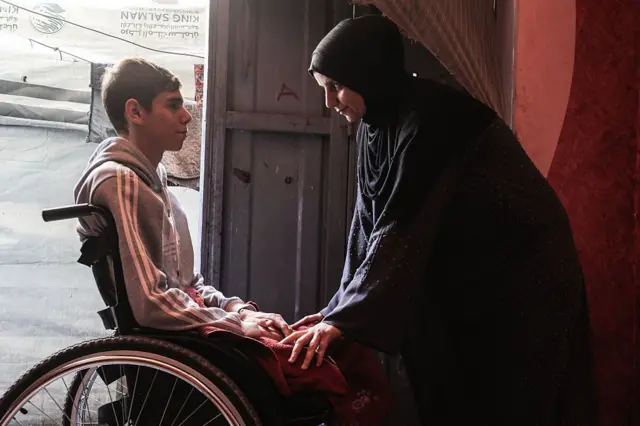 أحد الفتية مبتور الساقين من ضحايا الحرب في غزة يجلس على كرسي متحرك وبمواجهته أمه التي تعتني به بعد أن أصبح معوقا