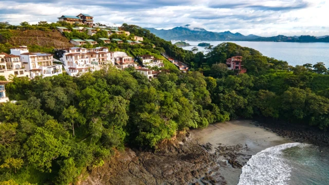 Na porośniętym drzewami wzgórzu stoją rzędy domów o białych i żółtych ścianach. Na pierwszym planie widoczny brzeg morza. W tle zatoka i kolejne pasma wzgórz.