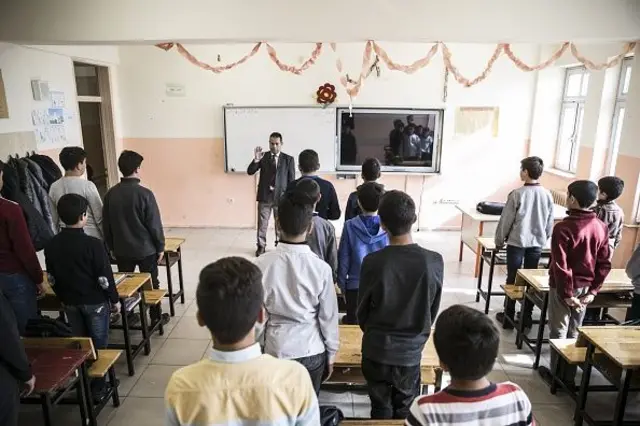 Arkası dönük ayakta öğrencilerin olduğu sınıfta öğretmen tahtanın önünde, oturun işareti yapıyor. 