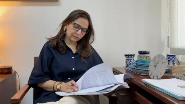 دکتر نبیهه فاطمه، جامعه‌شناس پاکستانی در پهلوی میز نشسته، دیک دست کاغذ و در دست دیگر قلم دارد و به کاغذهای که در دست دارد نگاه می‌کند. 