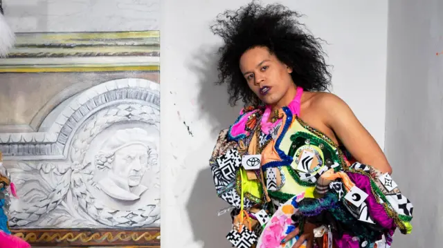 Quilla, con un atuendo colorido, de pie en una habitación, pero convertida en una obra de arte, mira seriamente a la cámara.