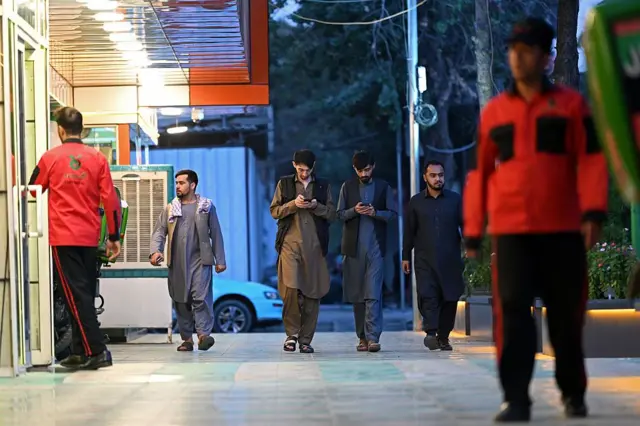 په عکس کې په نارنجي رنګه کمیس او تورو پرتوګونو کې د یوه رستورانت ګارسونان برېښي او شا ته ځينې ځوانان روان دي چې د دوه یې خپلو موبایلونو ته ځير دي.